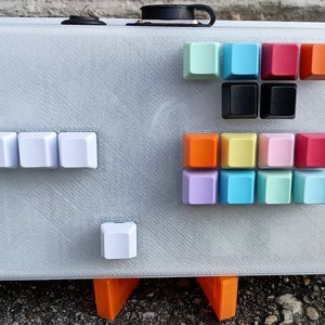 May include: A white plastic keyboard with a variety of colored keycaps. The keycaps are arranged in rows and columns, and they include colors such as red, orange, yellow, green, blue, and purple. The keyboard is sitting on a white surface.