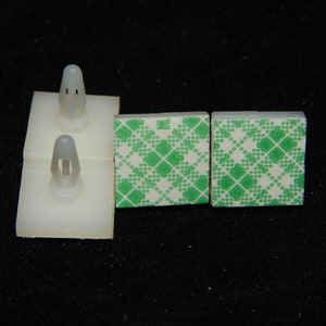 May include: A close-up shot of several white plastic mounting clips with gray push pins, and two small, square adhesive pads with a green and white diamond pattern. The items are on a black surface.