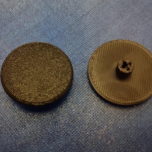 May include: Two black, round buttons. One button has a textured surface, while the other has a ribbed design and a four-hole shank. The buttons are likely for clothing or crafts.