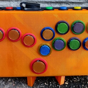 May include: Orange arcade button box with a variety of colored buttons. The box has a black strap and a black clip on the top. The box is sitting on a black surface.