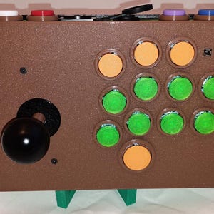 May include: A brown arcade cabinet with a joystick and 12 buttons. The buttons are arranged in a grid pattern with orange, green, and black buttons. The cabinet is sitting on two green stands.