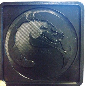 Pu&ograve; includere: Un sottobicchiere quadrato nero con un simbolo di drago nero e grigio in rilievo 3D al centro.
