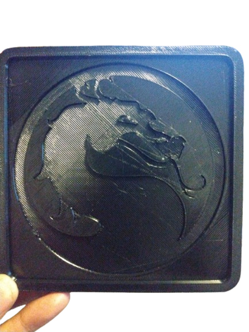 Pu&ograve; includere: Un sottobicchiere quadrato nero con un simbolo di drago nero e grigio in rilievo 3D al centro.