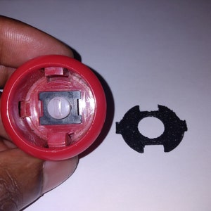 Puede incluir: Un bot&oacute;n de pl&aacute;stico rojo con una c&uacute;pula de pl&aacute;stico transparente y un interruptor negro en el interior. El bot&oacute;n se sostiene en una mano. Tambi&eacute;n se muestra un anillo de espuma negro.