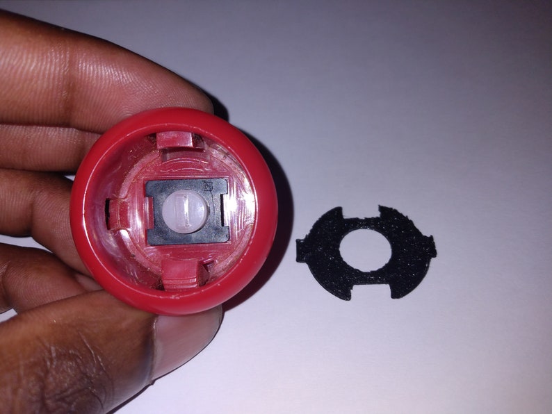 Puede incluir: Un bot&oacute;n de pl&aacute;stico rojo con una c&uacute;pula de pl&aacute;stico transparente y un interruptor negro en el interior. El bot&oacute;n se sostiene en una mano. Tambi&eacute;n se muestra un anillo de espuma negro.