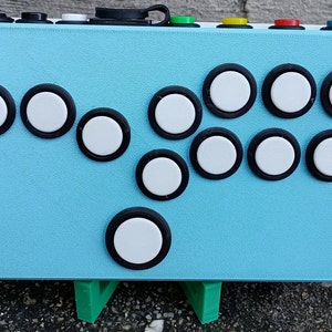 May include: A light blue electronic device with 11 white buttons with black borders. The device is on a green stand.