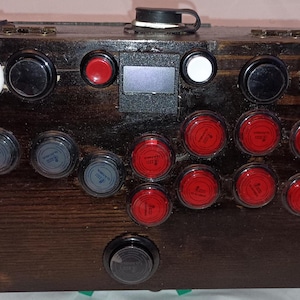 May include: A dark stained wooden box with various buttons and controls. The box features a combination of black, red, white, and gray buttons, along with a small screen and a black handle on top. The design suggests a control panel or gaming device.
