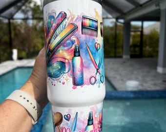 Puede incluir: Un vaso de acero inoxidable blanco con un diseño colorido que presenta herramientas para peinar el cabello, incluyendo tijeras, un cepillo para el cabello, laca para el cabello y una plancha para el cabello. El vaso tiene una tapa y un nombre personalizado, "Morgan", impreso en el lateral.