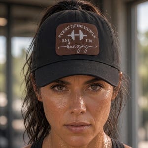 May include: A black baseball cap featuring a rectangular brown patch on the front. The patch displays a white graphic of a barbell and the text "EVERYTHING HURTS AND I'M hungry" in a combination of block and script lettering.