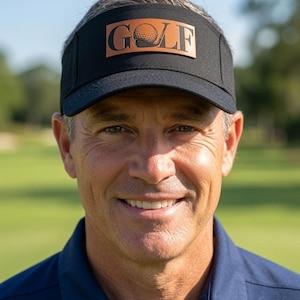 May include: A black golf visor featuring a rectangular brown leather patch on the front. The patch is embossed with the word GOLF, where the letter O is replaced by a textured golf ball graphic. The visor has a structured brim and crown.