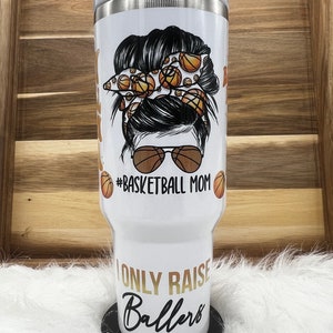 May include: White stainless steel tumbler with a black and white illustration of a woman with a messy bun decorated with basketballs. The text "I only raise ballers" is printed in gold on the tumbler. The text "#basketball mom" is printed in black below the illustration.
