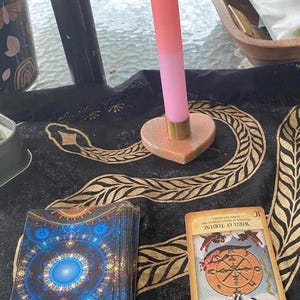 May include: A two-tone pink candle in a heart-shaped holder with a gold base. A deck of tarot cards with a blue and gold design is next to a single tarot card with the Wheel of Fortune. The items are on a black cloth with a gold pattern.