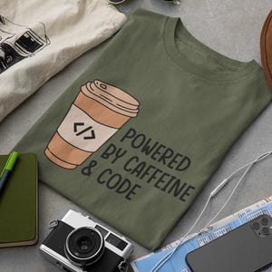 May include: An olive green t-shirt with a coffee cup graphic and the text "POWERED BY CAFFEINE & CODE." The image also includes a camera, notebook, hat, sunglasses, and a phone.