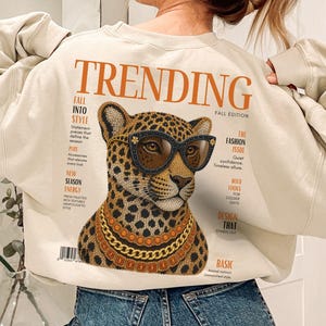 Könnte beinhalten: Ein beiges Sweatshirt mit Rundhalsausschnitt und einem großen Grafikdruck auf der Rückseite. Das Motiv zeigt einen Leoparden mit schwarz umrandeter Sonnenbrille und Goldketten im Stil eines Magazin-Covers mit dem Text TRENDING und FALL EDITION.