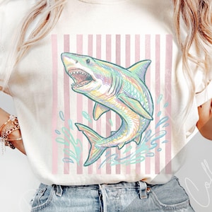 May include: Cream-colored t-shirt featuring a colorful shark illustration with open mouth and water splashes. The shark is set against a pink and white striped background. The design is a playful and artistic representation of marine life.