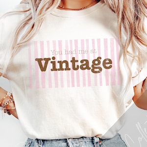 May include: A cream-coloured sweatshirt featuring the phrase "You had me at Vintage" in brown, set against a pink and white striped design. The sweatshirt is worn with denim shorts.