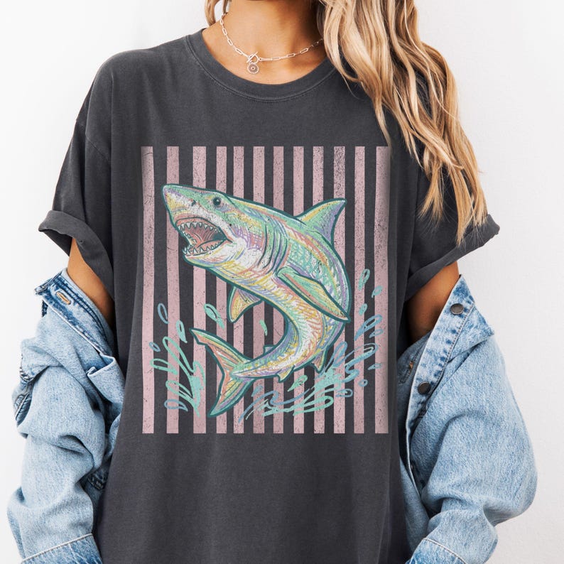 May include: Dark gray t-shirt featuring a colorful shark illustration with an open mouth, set against a pink and white striped background. The shark is detailed with shades of blue, green, and yellow, with water splashes.