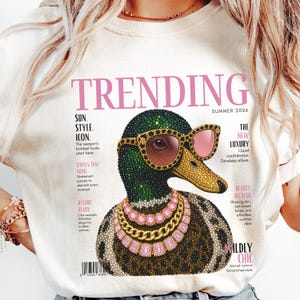 May include: A cream-colored t-shirt featuring a graphic of a mallard duck wearing pink-tinted sunglasses and layered gold and pink gemstone necklaces. The design mimics a magazine cover with the text TRENDING, SUMMER 2026, and various headlines.