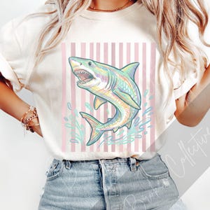 May include: Cream-colored t-shirt featuring a colorful shark illustration with open jaws and water splashes. The shark design has rainbow hues and is set against a pink and white striped background. The shirt is paired with denim shorts.