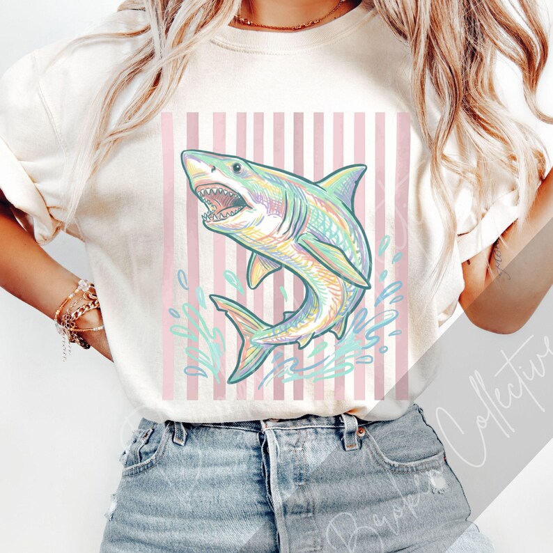 May include: Cream-colored t-shirt featuring a colorful shark illustration with open jaws and water splashes. The shark design has rainbow hues and is set against a pink and white striped background. The shirt is paired with denim shorts.