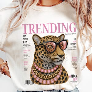 Könnte beinhalten: Ein cremefarbenes T-Shirt mit einer Grafik eines Leoparden, der eine schwarze Sonnenbrille und pinkfarbene Edelsteinketten trägt. Das Design ähnelt einem Magazin-Cover mit dem Text TRENDING, SUMMER 2026 und verschiedenen Mode-Slogans.