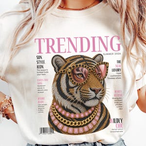 May include: A cream-colored t-shirt featuring a graphic print of a tiger wearing pink-tinted sunglasses and layered gold and pink gemstone necklaces. The design mimics a magazine cover with the text TRENDING, SUMMER 2026, and various headlines.