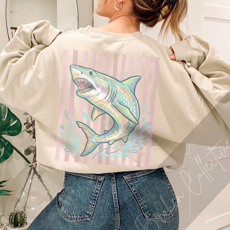 May include: Beige sweatshirt featuring a colorful shark graphic on the back. The shark is depicted with open jaws, surrounded by water splashes, and set against a backdrop of pink and white vertical stripes. The sweatshirt has long sleeves and a crew neck.
