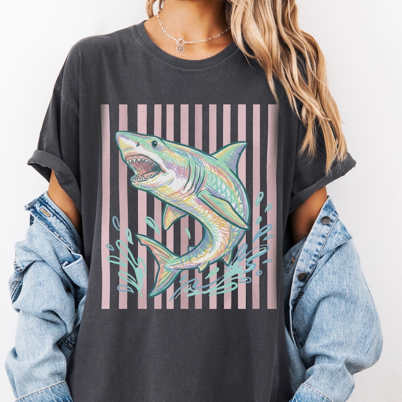 May include: Dark gray t-shirt featuring a colorful shark graphic. The shark is depicted with open jaws, surrounded by water splashes, set against a pink and black striped background. The shirt has rolled-up sleeves.