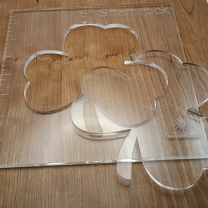 May include: A clear acrylic stencil of a 6-inch shamrock. The stencil is on a wooden surface.
