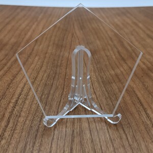 May include: A clear acrylic display stand with a triangular base. The stand is designed to hold a piece of art or a photograph.