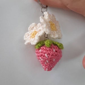 以下が含まれることがあります： 銀色のキーリングに、2つの白い編み花の付いた、編み物のイチゴのキーホルダーです。