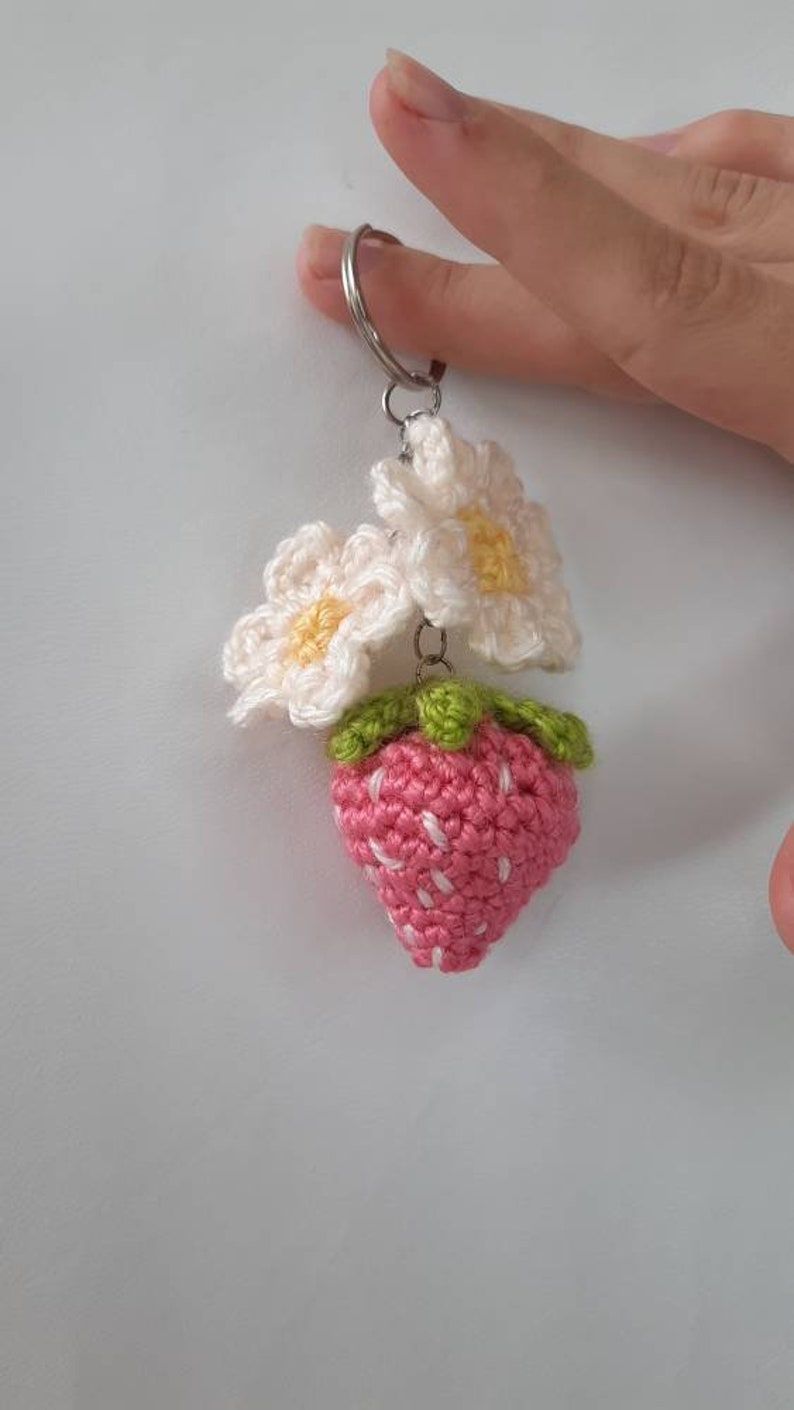 以下が含まれることがあります： 銀色のキーリングに、2つの白い編み花の付いた、編み物のイチゴのキーホルダーです。