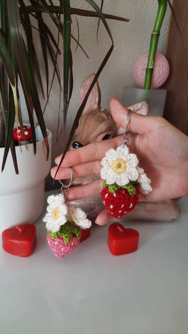 以下が含まれることがあります： 白い花が付いた、手編みのイチゴのキーホルダーが2つ。イチゴは赤く、緑の葉と白い点々があります。キーホルダーは銀色のキーリングに付いています。