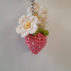 以下が含まれることがあります： 上部に白い花が2つ付いた、かぎ針編みで作ったイチゴのキーホルダー。キーホルダーは銀色のキーリングに付いています。