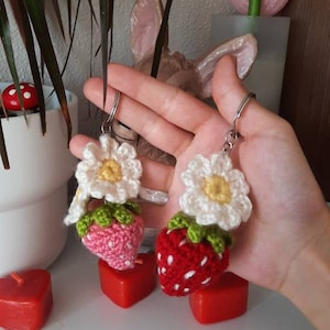 以下が含まれることがあります： 白い花が付いた、手編みのイチゴのキーホルダーが2つ。イチゴは赤とピンクで、緑の葉が付いています。キーホルダーは銀色のキーリングに付いています。