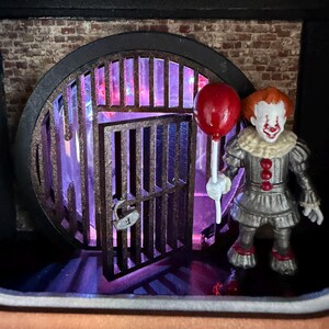 May include: A miniature diorama featuring a clown figurine with orange hair, white face paint, and a grey ruffled costume, holding a single red balloon. The figure stands before a circular black metal gate with a padlock, set against a brick wall.