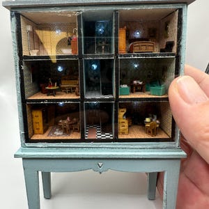 May include: A miniature dollhouse with nine rooms, each meticulously furnished. The dollhouse has a light blue base and a gray roof. The rooms are lit with small lights, showcasing detailed interior designs, including a bathroom, bedroom, and living room.