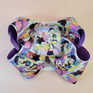 May include: A large hair bow made of white grosgrain ribbon featuring a repeating anime-style character print in shades of pink, purple, yellow, and blue. A central glittery rectangular charm displays the text HUNTRX and three anime characters.