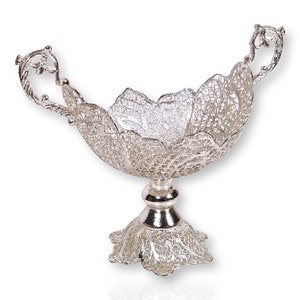Puede incluir: Un cuenco decorativo de plata con un diseño intrincado y de encaje. El cuenco tiene dos asas y se asienta sobre una base de pedestal.
