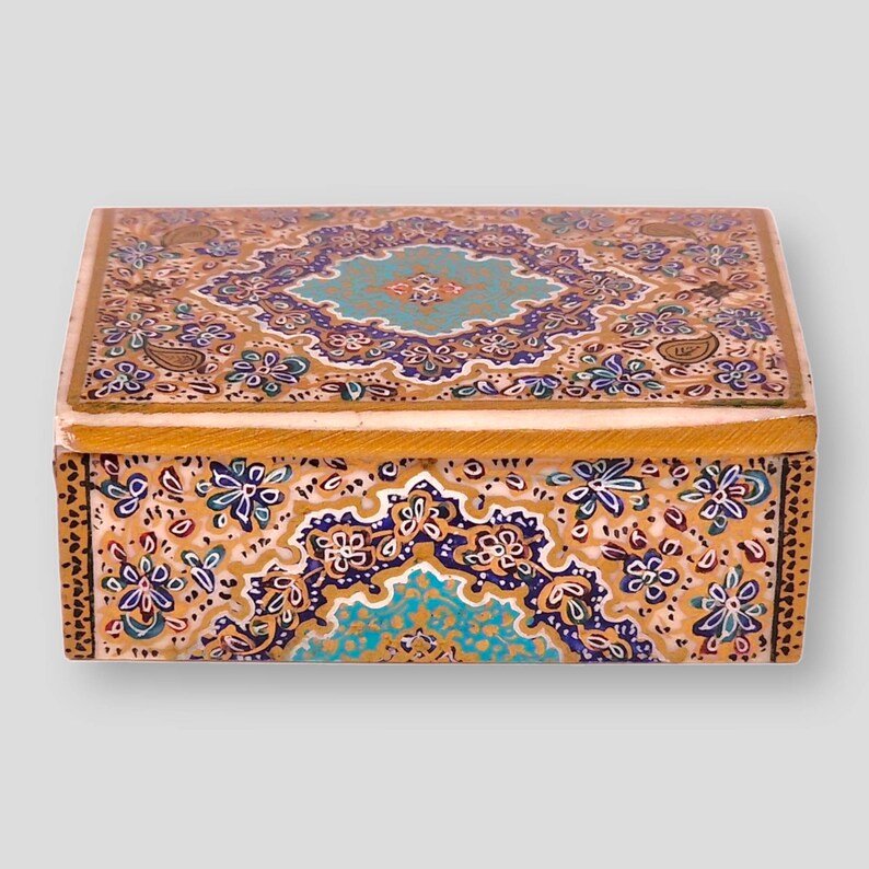 May include: A small wooden box with a decorative floral design. The box is painted in shades of gold, blue, green, and white. The design features intricate paisley patterns and floral motifs.