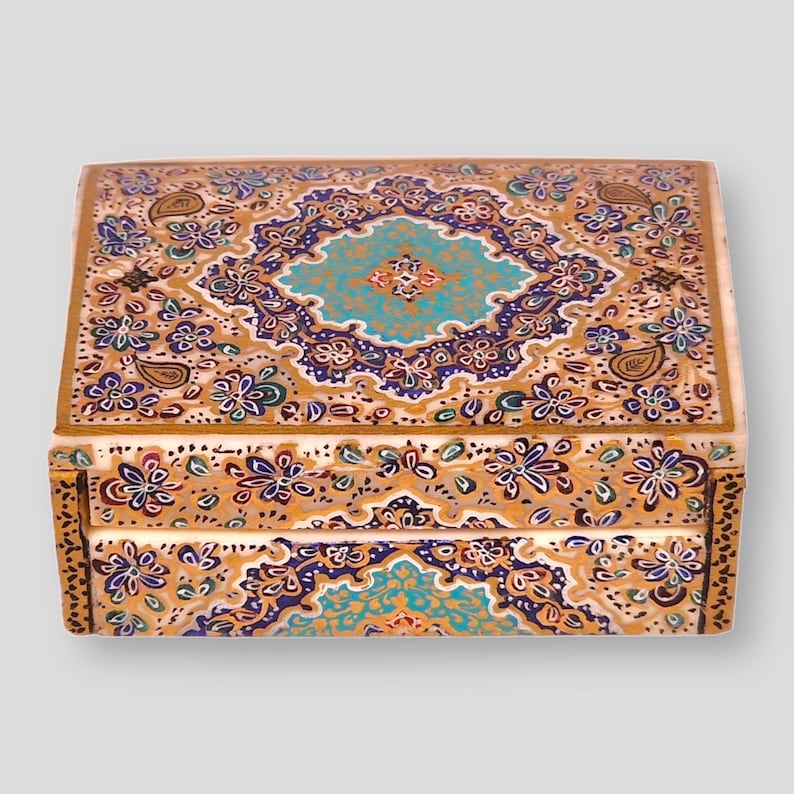 May include: A small, intricately decorated wooden box with a gold background and a blue and purple floral pattern. The box has a hinged lid and is perfect for storing small items.
