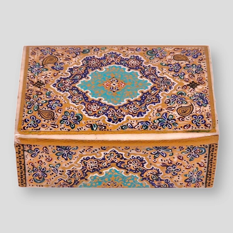 May include: A small wooden box with a decorative design. The box is painted with a gold background and intricate patterns in blue, green, and white. The lid of the box is closed.