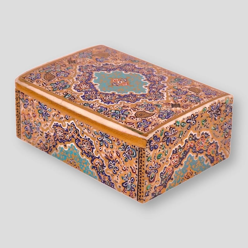 May include: A small wooden box with a decorative lid. The lid is painted with intricate floral patterns in shades of gold, blue, and purple. The box is a traditional style and would be a beautiful addition to any home.
