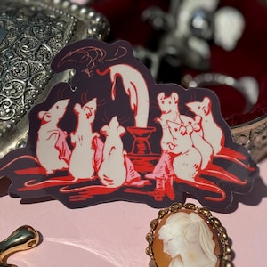 May include: A sticker depicting white mice around a candle, set against a dark purple backdrop with red details. A silver jewellery box and a cameo brooch are also visible, all on a pink surface.
