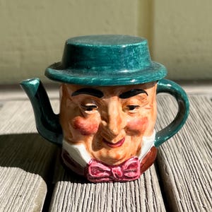 Puede incluir: Una tetera de cerámica verde en forma de cabeza de hombre con un sombrero verde y una pajarita roja.