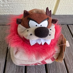 May include: A plush toy of Taz the Tasmanian Devil from the Looney Tunes cartoon. The toy is brown and has a red fur collar. The toy is wearing a tag that says "Taz"