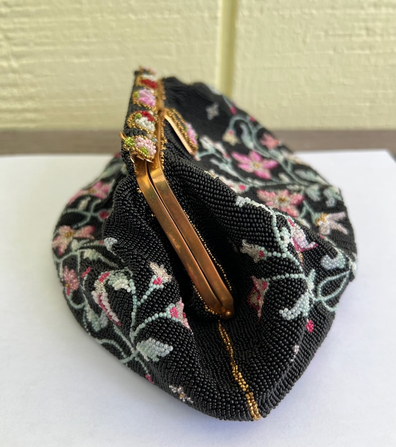 May include: A black beaded evening bag with a floral design in pink, white, and green. The bag has a gold clasp and a beaded trim.