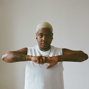 May include: A person wearing a white sleeveless shirt and silver jewelry, including a chain necklace and bracelet. The person has tattoos on their arms and is making a fist with both hands. They have short, wavy, blonde hair and are wearing earrings and a nose ring.
