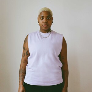 May include: A person wearing a sleeveless lavender shirt and black pants. The person has short, light-colored hair, multiple ear piercings, and a silver chain necklace. Tattoos are visible on both arms.