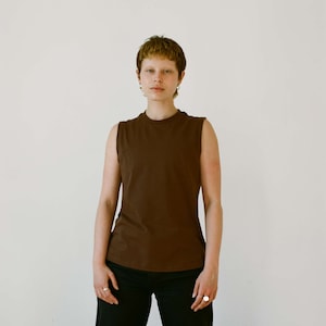 May include: A sleeveless, dark brown top paired with black pants. The top has a crew neck and a fitted silhouette. The model is standing in front of a plain white background. The outfit is simple and casual.